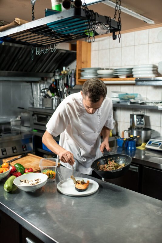 young male chef white uniform putting roasted vegetables piece fried salmon while preparing tasty meal client restaurant